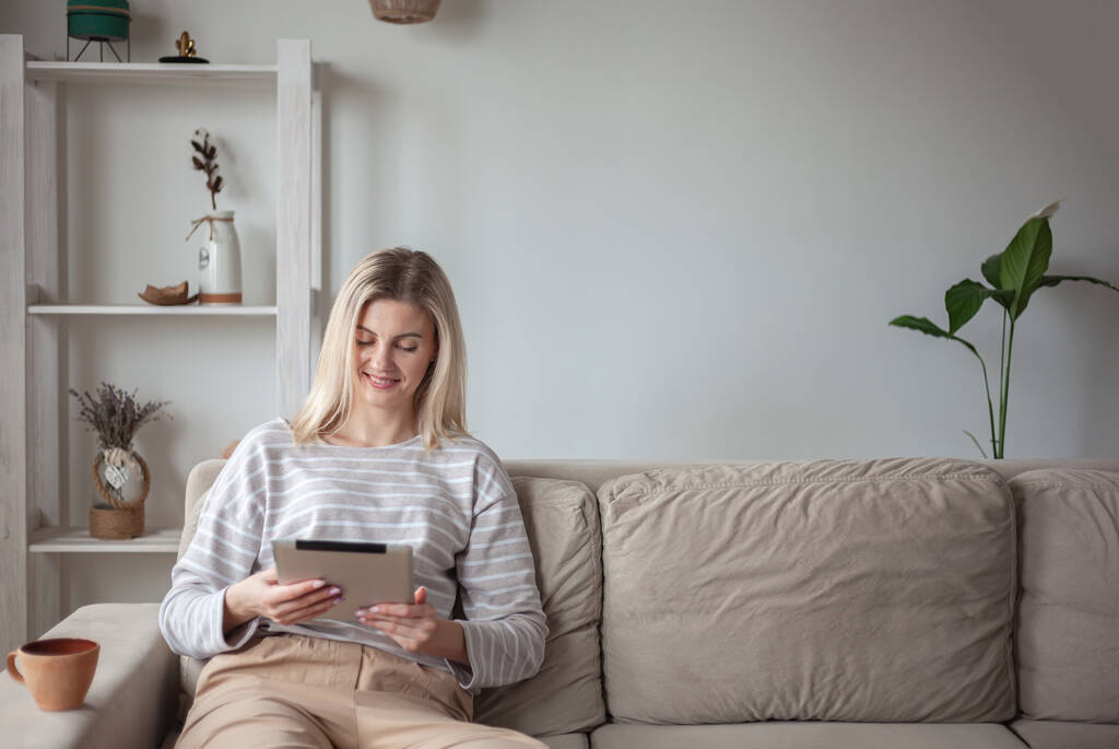 Молода жінка використовує планшет, сидить на дивані вдома і п'є каву
 - Фото, зображення