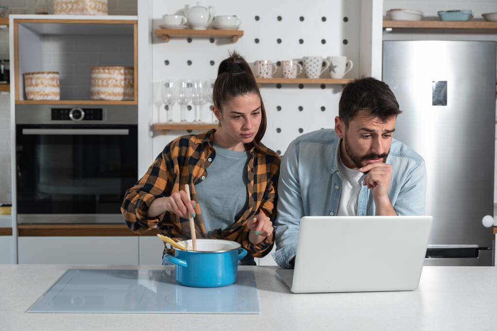 Νεαρό ζευγάρι στέκεται στην κουζίνα και βλέποντας μια online συνταγή στο διαδίκτυο σε ένα φορητό υπολογιστή, ενώ μαγειρεύουν ένα νέο πιάτο σε μια κατσαρόλα με ζυμαρικά και η γυναίκα κρατά μια κουζίνα και ανάδευση  - Φωτογραφία, εικόνα