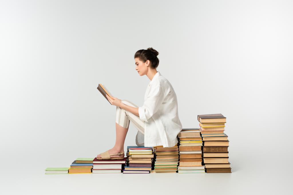 Seitenansicht einer jungen Frau, die auf einem Bücherstapel sitzt, während sie auf Weiß liest  - Foto, Bild