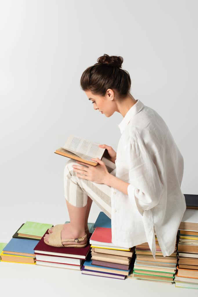 Genç esmer kadın, beyazın üzerinde izole bir şekilde kitap yığını üzerinde otururken kitap okuyor.  - Fotoğraf, Görsel