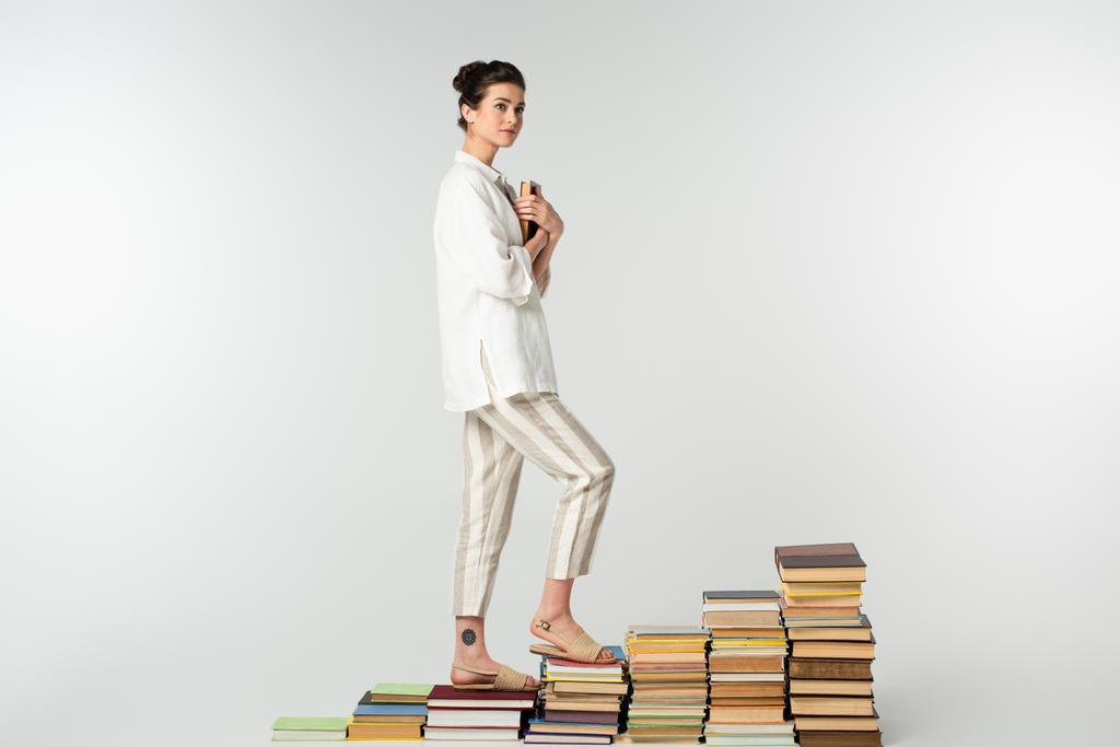 full length of young tattooed woman standing on pile of books isolated on white  - Photo, Image