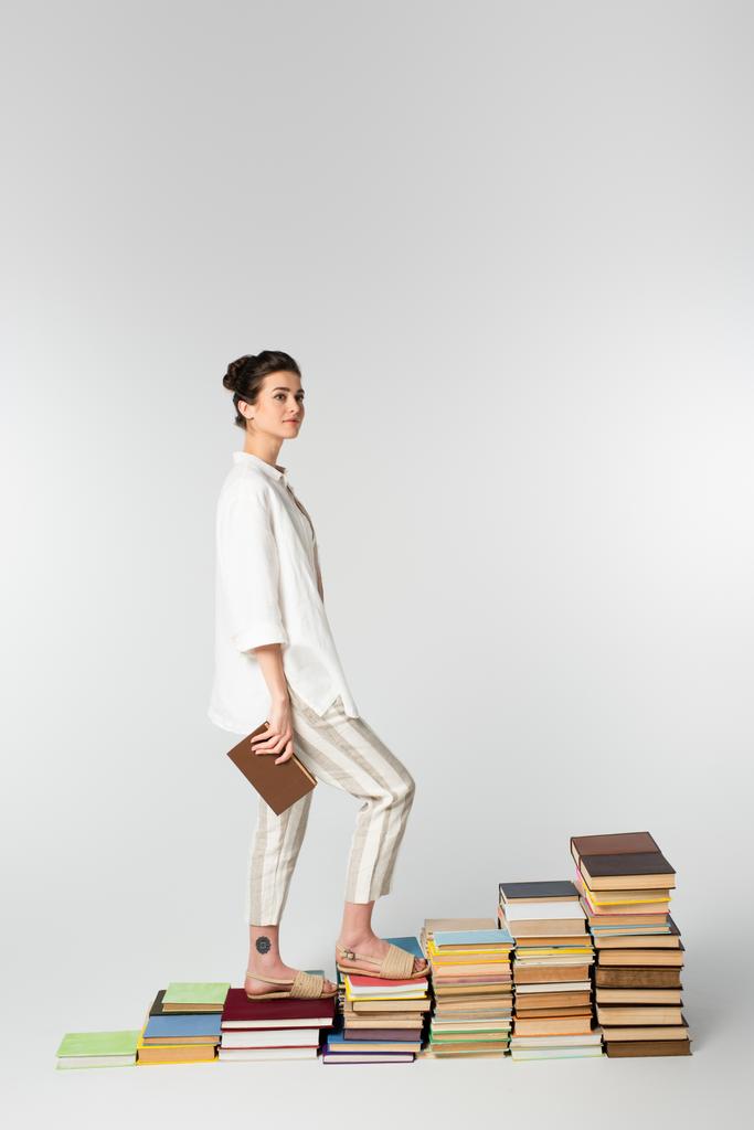 полная длина молодой женщины с татуировкой, стоящей на куче книг, изолированных на белом  - Фото, изображение