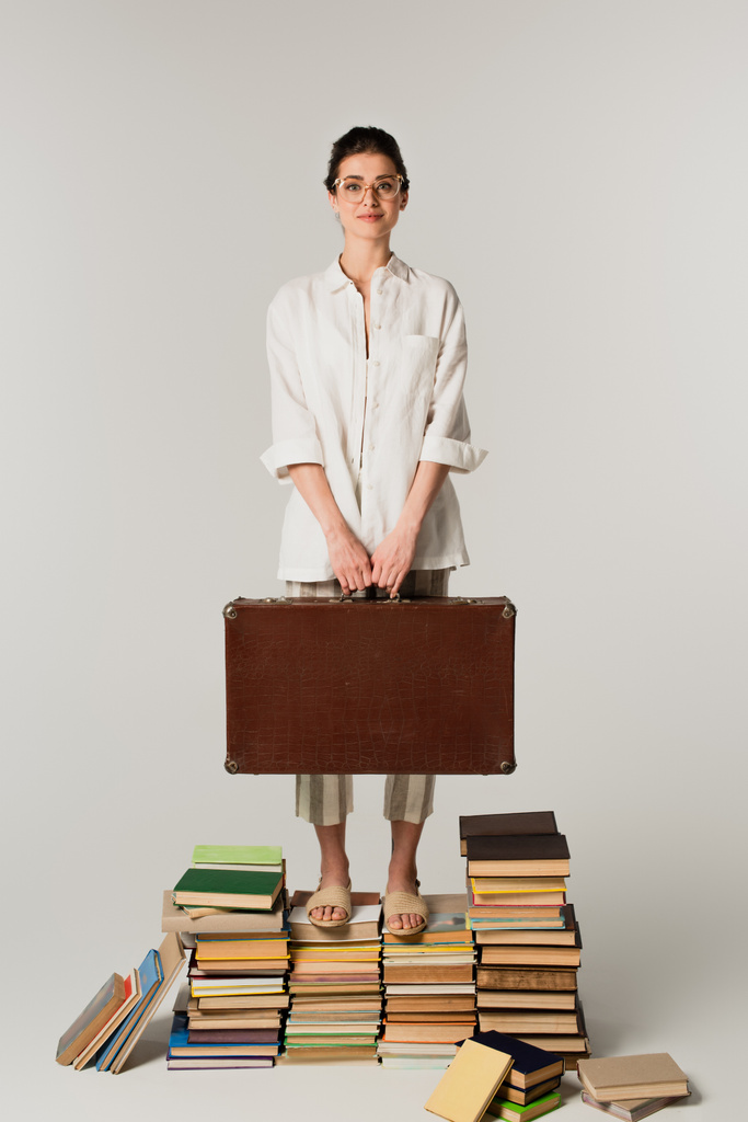 pełna długość zadowolonej młodej kobiety w okularach trzymającej walizkę stojąc na stosie książek odizolowanych na biało  - Zdjęcie, obraz