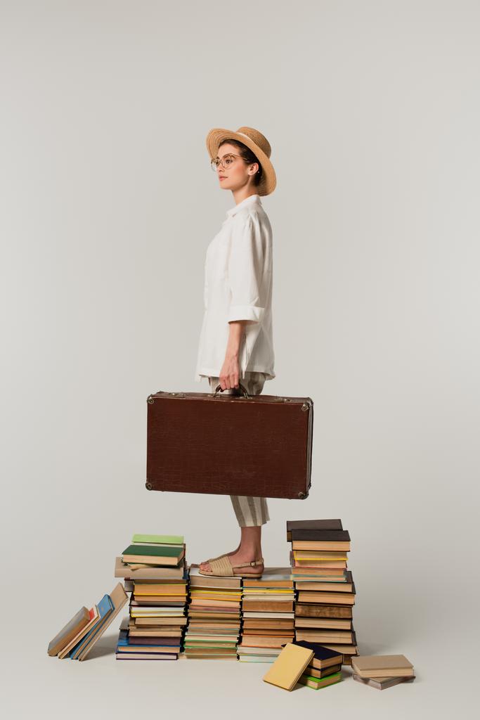 longitud completa de la mujer joven en sombrero de paja y gafas que sostienen la maleta mientras está de pie en la pila de libros aislados en blanco  - Foto, Imagen