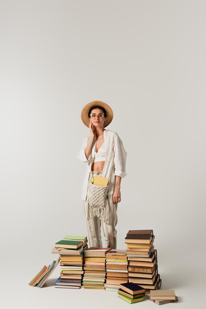 mujer joven complacida en sombrero de sol de pie cerca de pila de libros aislados en blanco - Foto, imagen