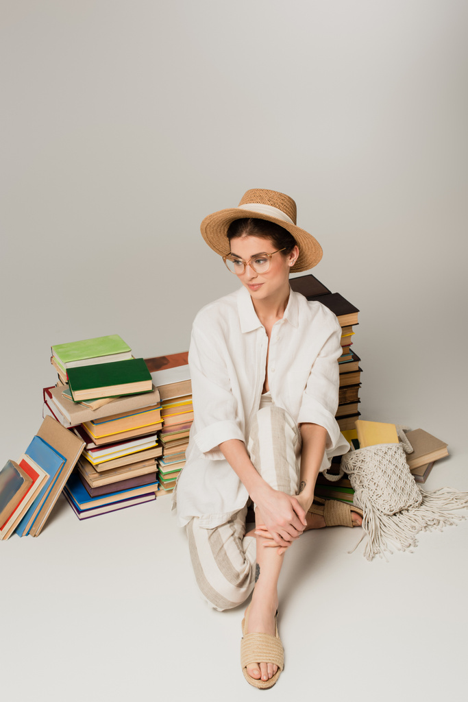 молода жінка в солом'яному капелюсі і окулярах, що сидить біля купи книг на білому
 - Фото, зображення