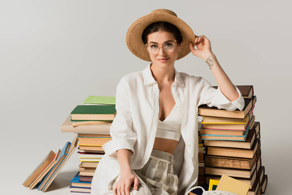 zufriedene und tätowierte Frau mit Strohhut und Brille, die neben einem Bücherstapel auf weißem Papier sitzt - Foto, Bild