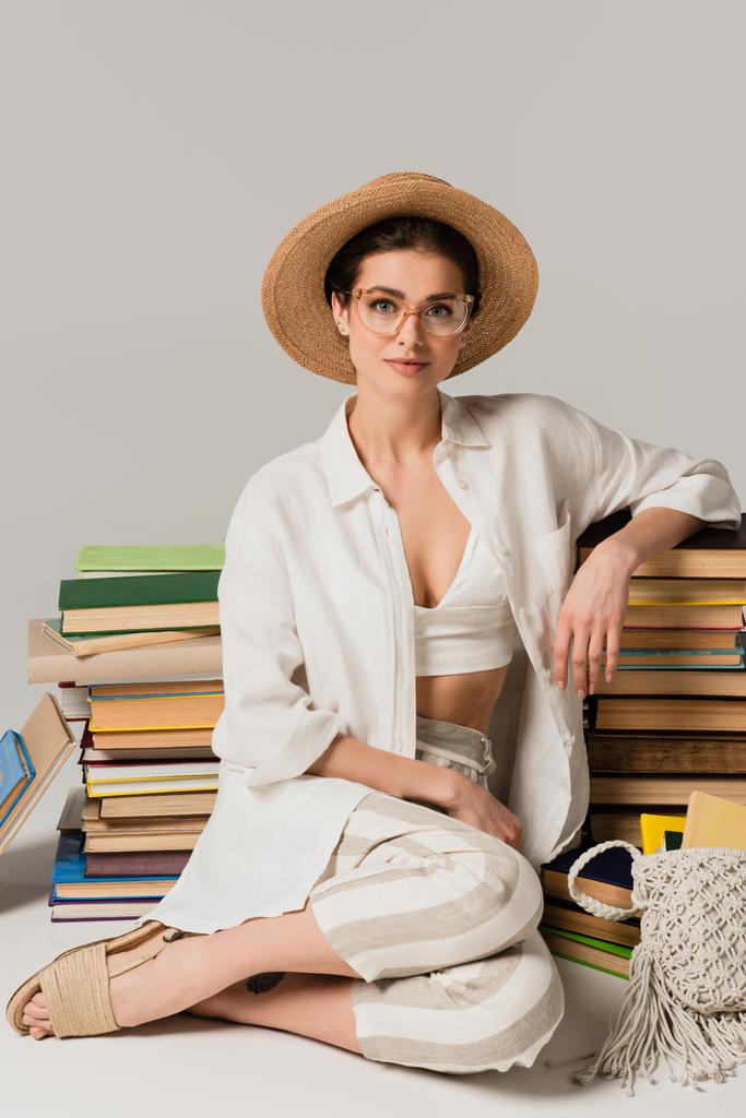 tevreden vrouw in stro hoed en bril zitten in de buurt stapel boeken op wit - Foto, afbeelding