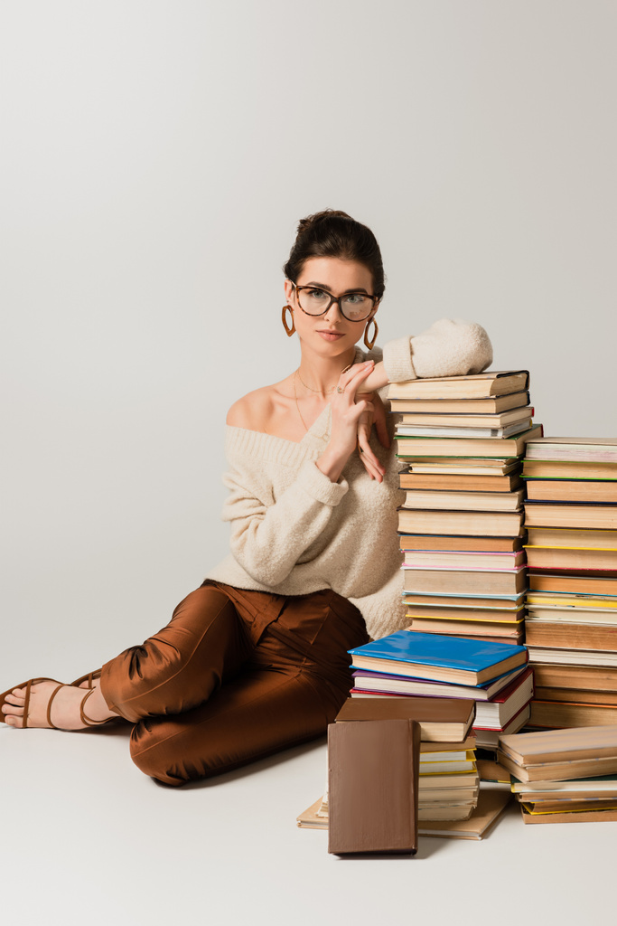 piena lunghezza della giovane donna in occhiali e orecchini appoggiati su pila di libri su bianco - Foto, immagini