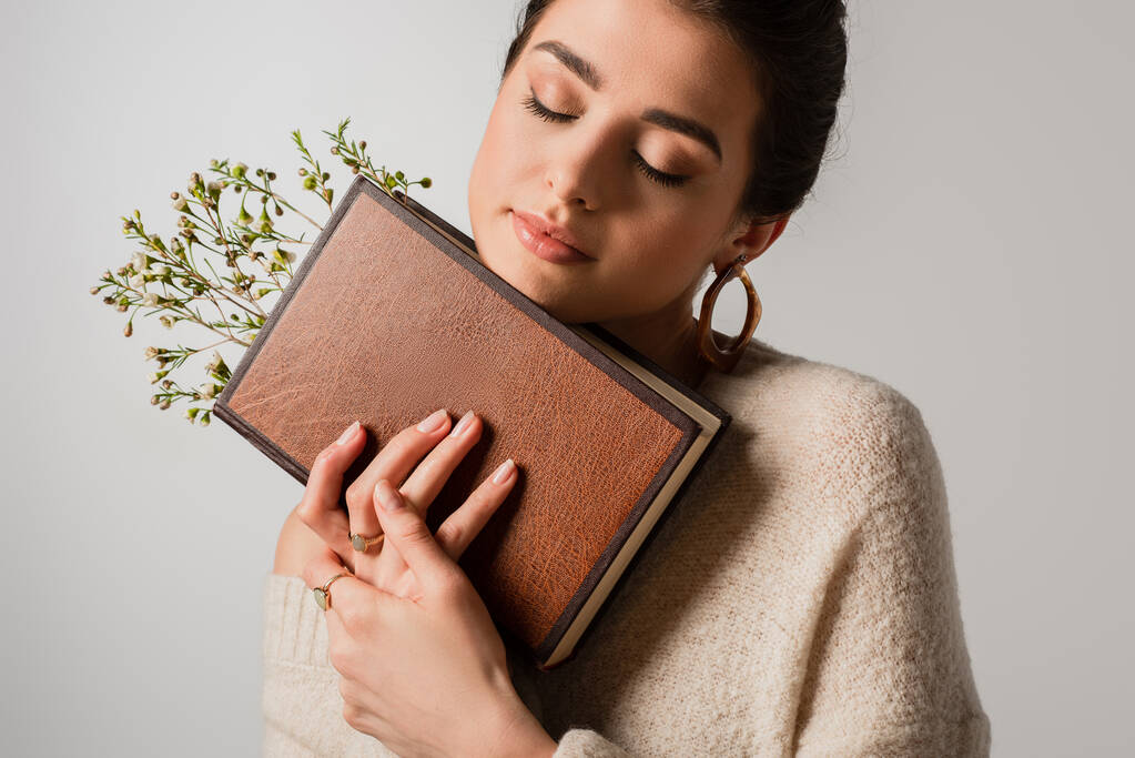 jovem com olhos fechados segurando livro com flores silvestres isolado em cinza - Foto, Imagem