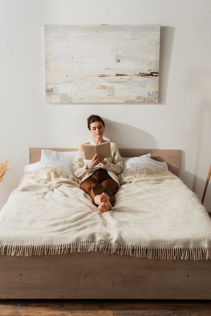 barefoot young woman reading book while resting on bed at home - Photo, Image