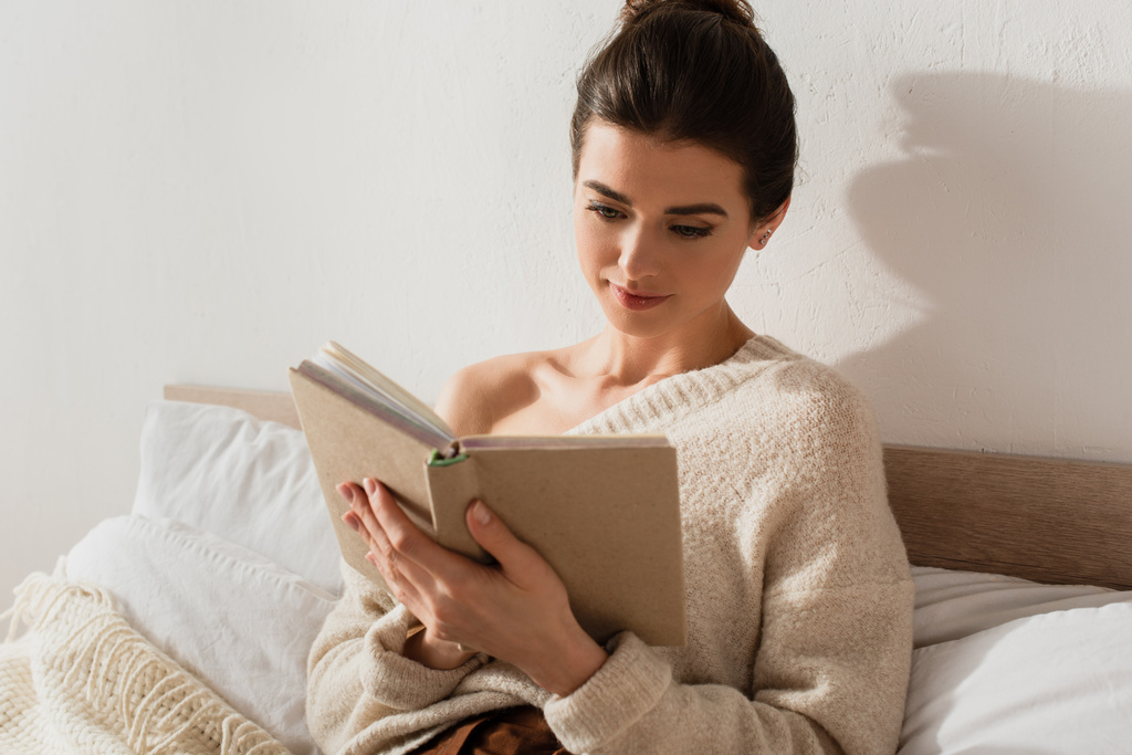hezká mladá žena čtení knihy, zatímco odpočívá na posteli doma - Fotografie, Obrázek