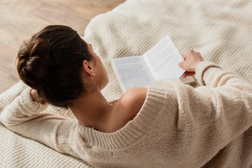 πίσω όψη της νεαρής μελαχρινής γυναίκας ανάγνωση βιβλίο, ενώ στηρίζεται στο κρεβάτι στο σπίτι - Φωτογραφία, εικόνα
