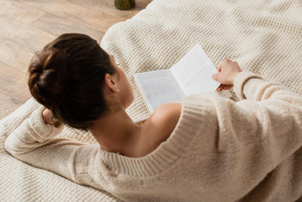 back view of young brunette woman in sweater reading book while resting on bed at home - Photo, Image