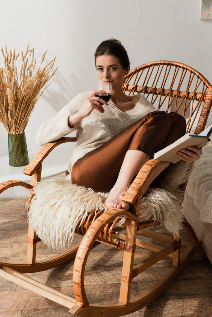 молода жінка сидить у плетеному кріслі з книгою та п'є червоне вино
 - Фото, зображення