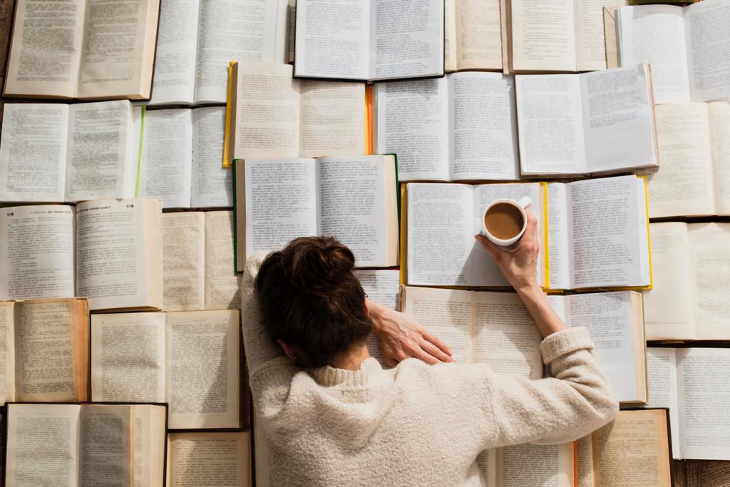 κορυφαία άποψη της κουρασμένης γυναίκας κοιμάται κοντά σε ανοιχτά βιβλία με φλιτζάνι καφέ - Φωτογραφία, εικόνα