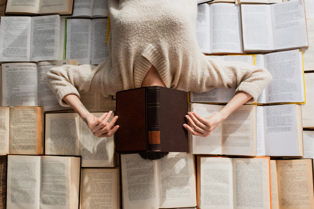 верхний вид татуированной женщины, скрывающей лицо, лежащей на груде открытых книг  - Фото, изображение
