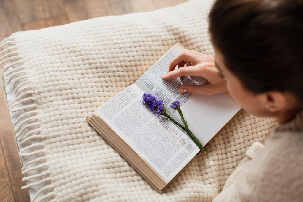 θέα υψηλής γωνίας του θολή νεαρή γυναίκα ανάγνωση βιβλίο με μωβ λουλούδι, ενώ στηρίζεται στο κρεβάτι στο σπίτι - Φωτογραφία, εικόνα