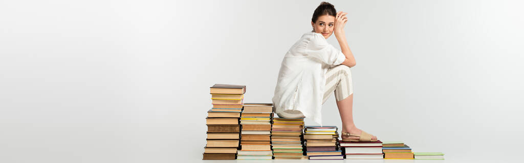 longitud completa de la mujer joven en sandalias sentadas en la pila de libros en blanco, bandera - Foto, imagen
