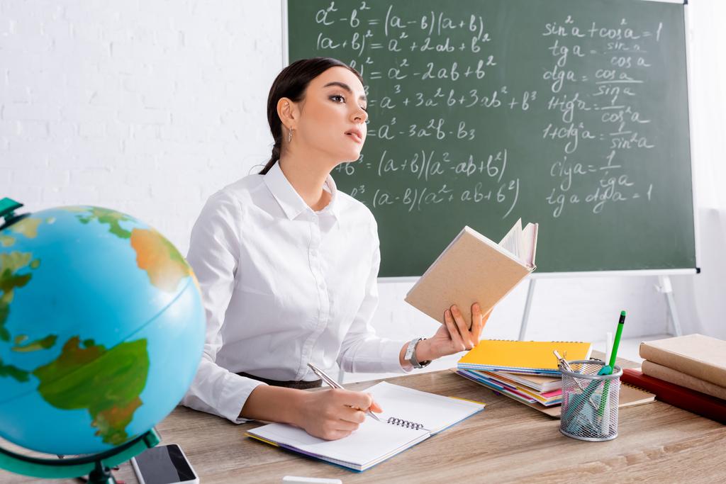 Teacher holding book and writing on notebook near globe in classroom  - Photo, Image