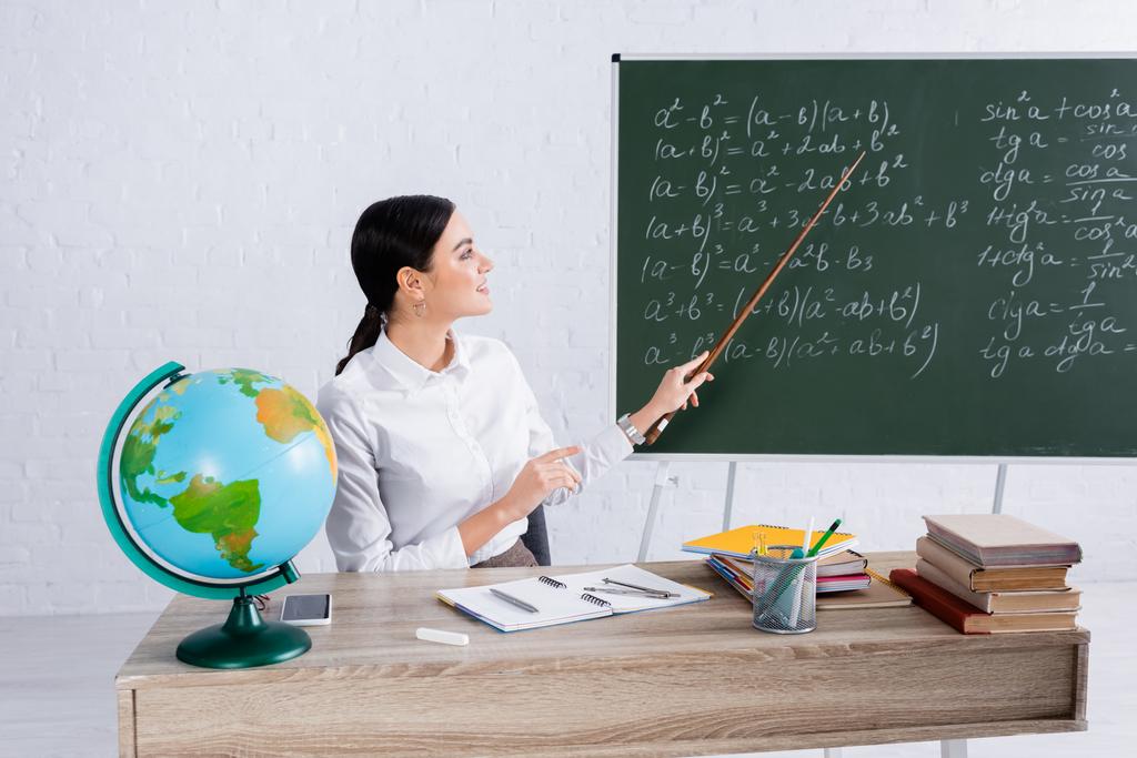 Gülümseyen öğretmen okulda kitapların ve dünyanın yakınındaki denklemlerle tahtayı işaret ediyor.  - Fotoğraf, Görsel