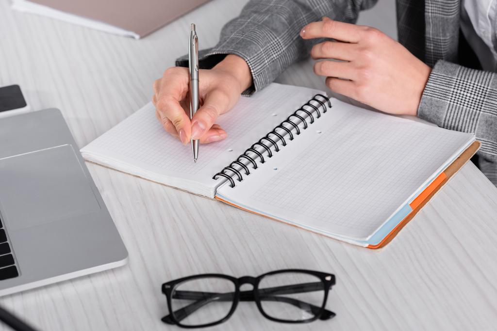 Vue recadrée d'une femme d'affaires écrivant sur un carnet près de lunettes et d'un ordinateur portable  - Photo, image