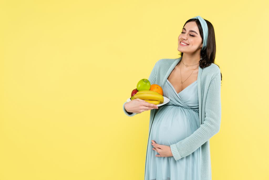 donna incinta sorridente che tiene il piatto con frutti maturi isolati sul giallo - Foto, immagini