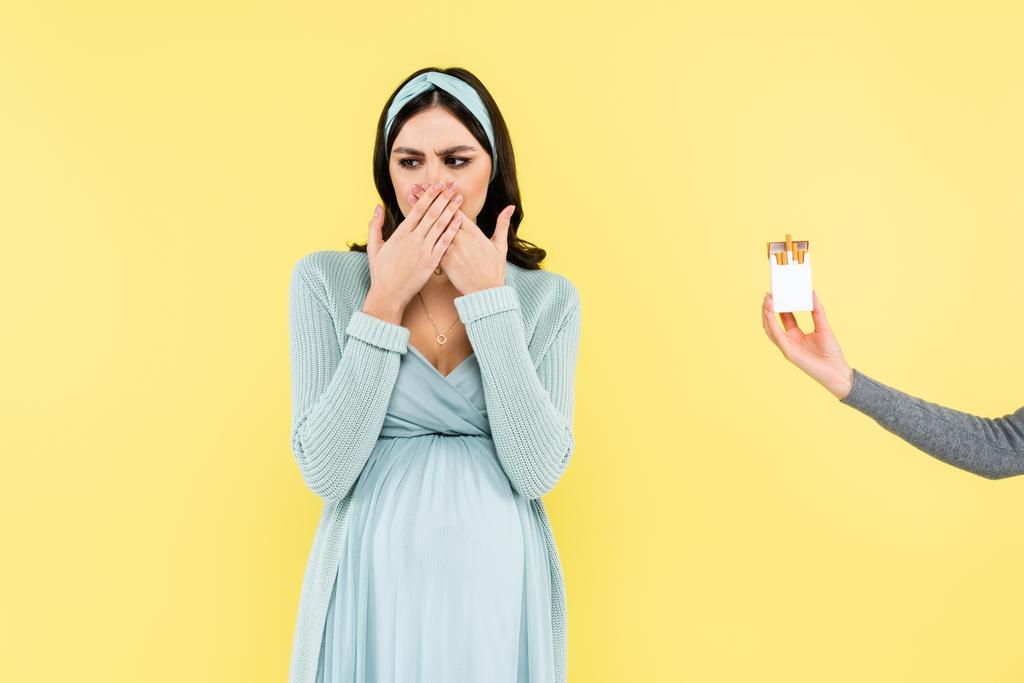 Schwangere empfindet Ekel und deckt Mund in der Nähe von Zigarettenschachtel isoliert auf gelb - Foto, Bild