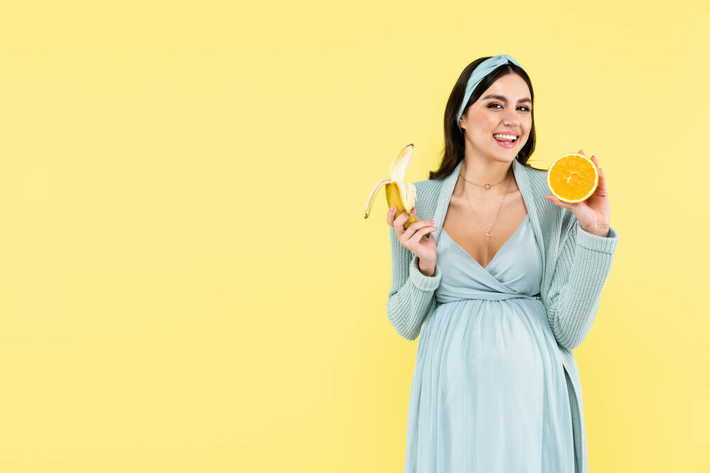 glücklich schwangere Frau lächelt in die Kamera, während sie Banane und saftige Orange isoliert auf gelb hält - Foto, Bild