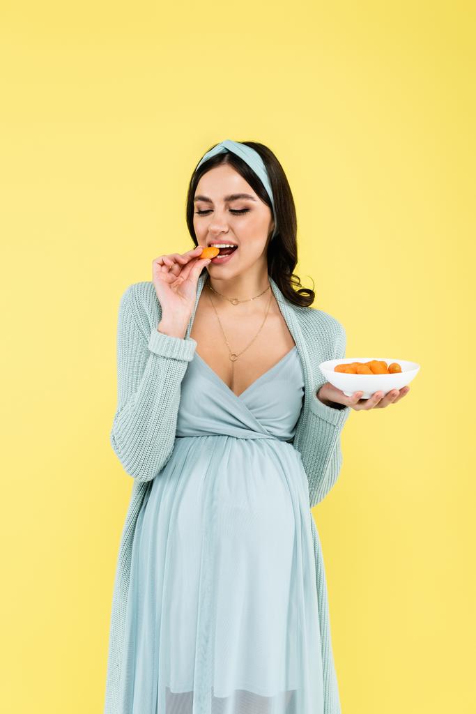 mladá těhotná žena jíst čerstvé mrkev izolované na žluté - Fotografie, Obrázek