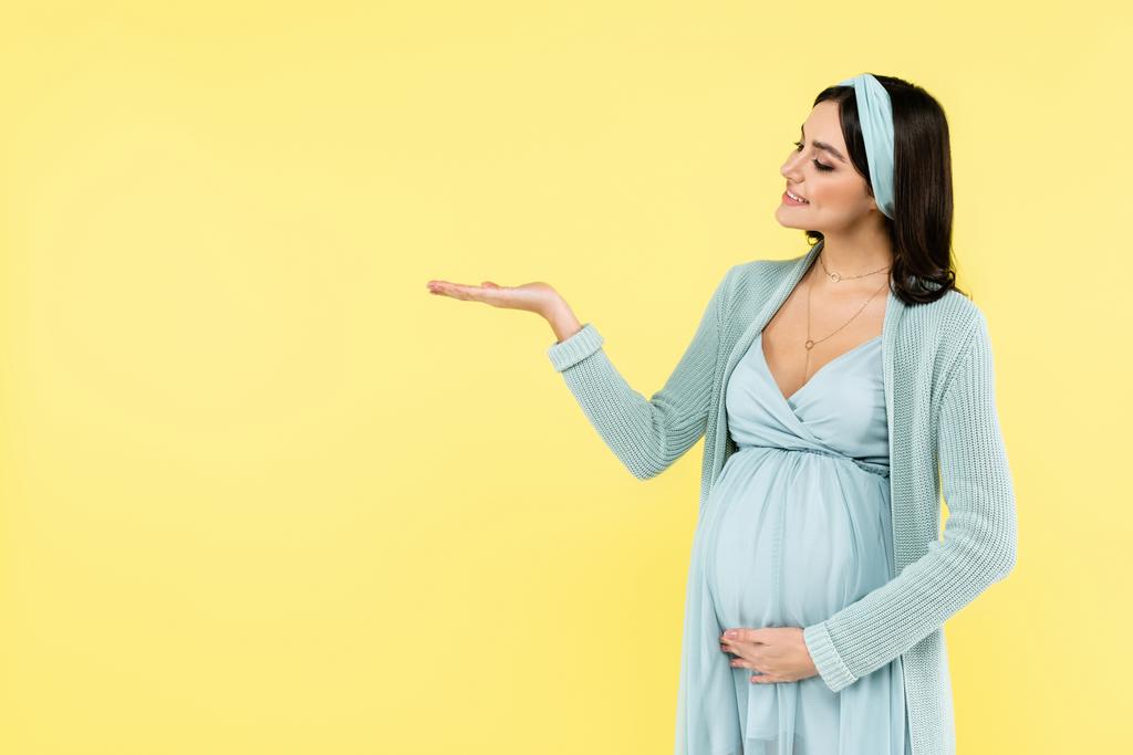 χαμογελαστή έγκυος γυναίκα δείχνει με το χέρι απομονώνονται σε κίτρινο - Φωτογραφία, εικόνα