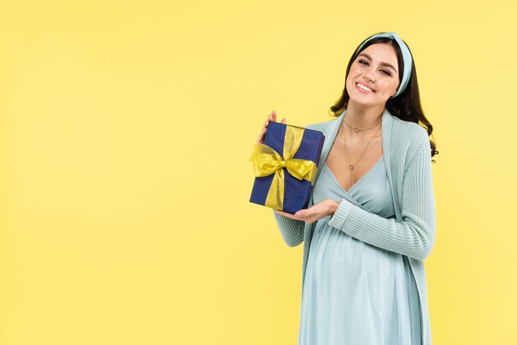 Neşeli hamile kadın kameraya gülümserken sarıda izole edilmiş hediyesini tutuyor. - Fotoğraf, Görsel