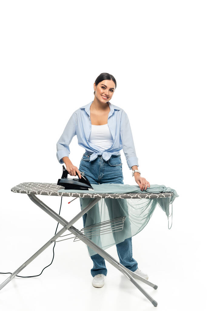 mujer alegre sonriendo a la cámara mientras plancha la ropa sobre fondo blanco - Foto, Imagen