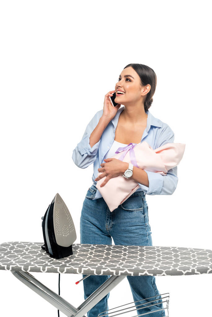 Радостная женщина с младенцем разговаривает на смартфоне рядом с гладильной доской изолированы на белом - Фото, изображение