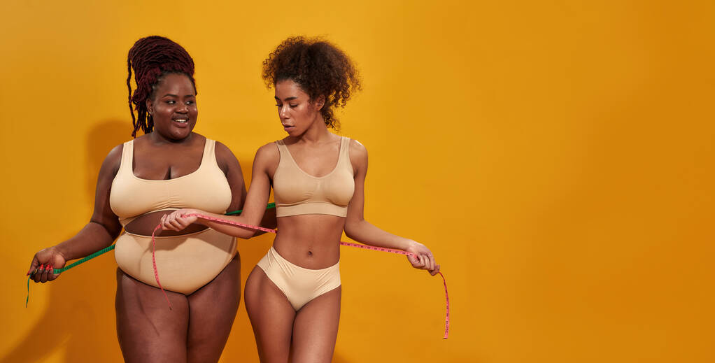 Студийный снимок двух африканских американок в нижнем белье с разной формой тела, держащих метрическую ленту, стоящих изолированно на оранжевом фоне - Фото, изображение