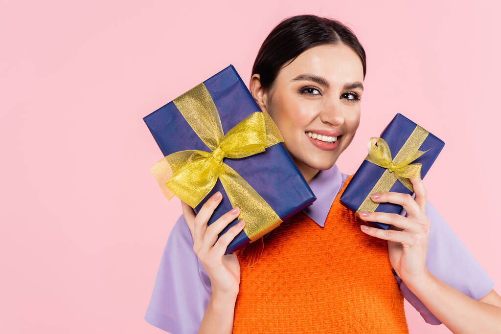 brunette femme souriant à la caméra tout en tenant des cadeaux isolés sur rose - Photo, image