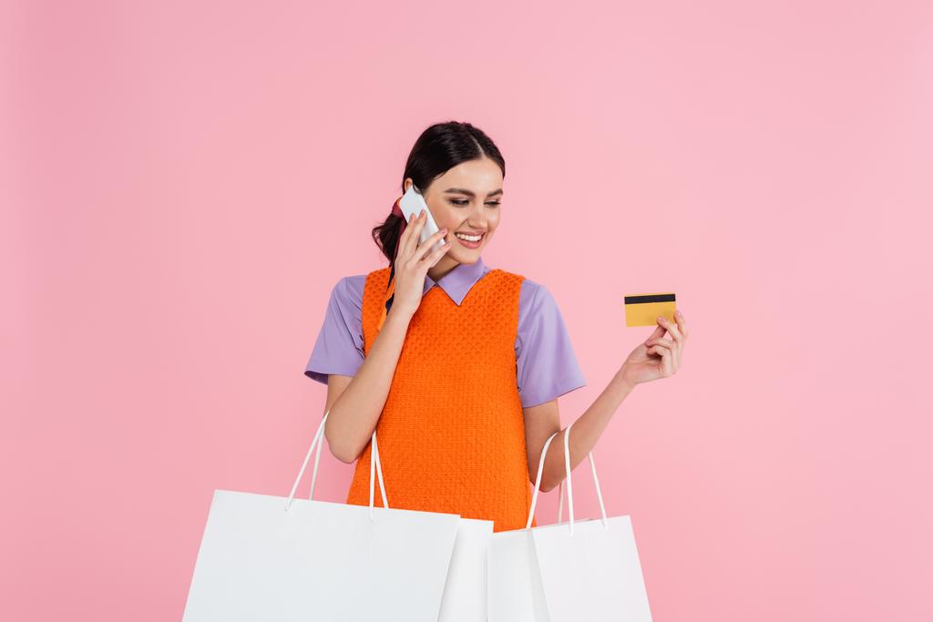 donna felice con carta di credito e shopping bag che parla su smartphone isolato su rosa - Foto, immagini