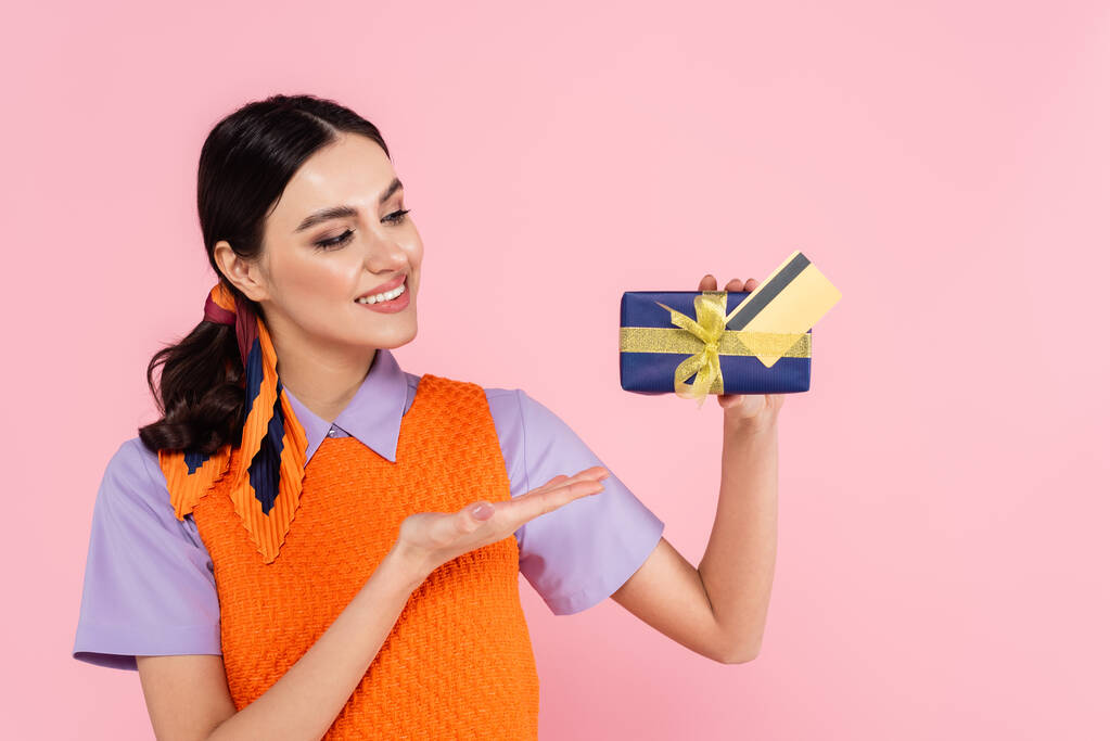 mujer alegre señalando caja de regalo y tarjeta de crédito aislada en rosa - Foto, imagen