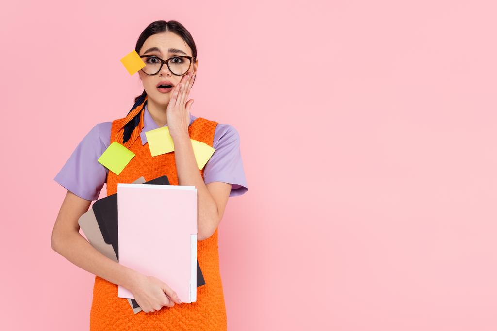 σοκαρισμένη επιχειρηματίας με κολλώδεις σημειώσεις για τα ρούχα και γυαλιά κρατώντας έγγραφα που απομονώνονται σε ροζ - Φωτογραφία, εικόνα
