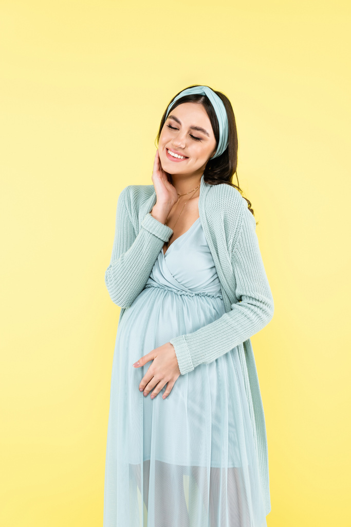 mulher grávida feliz tocando rosto e barriga enquanto sorrindo com os olhos fechados isolados no amarelo - Foto, Imagem