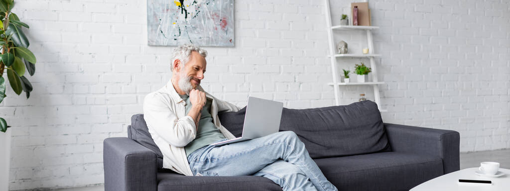 homme barbu regardant ordinateur portable assis sur le canapé, bannière - Photo, image