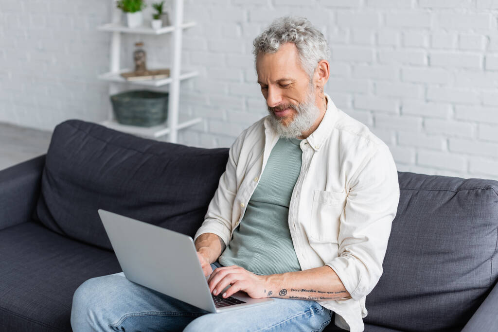 tetovált férfi használ laptop, miközben ül a kanapén otthon - Fotó, kép