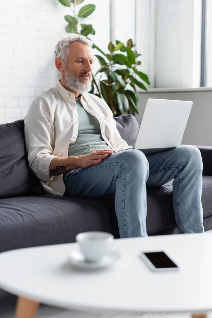 γενειοφόρος άντρας που χρησιμοποιεί φορητό υπολογιστή ενώ κάθεται στον καναπέ κοντά σε φλιτζάνι καφέ και smartphone με λευκή οθόνη σε θολή πρώτο πλάνο - Φωτογραφία, εικόνα