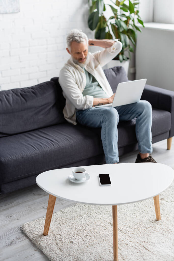 cup of coffee and smartphone with blank screen on coffee table near bearded man using laptop on blurred background  - Photo, Image