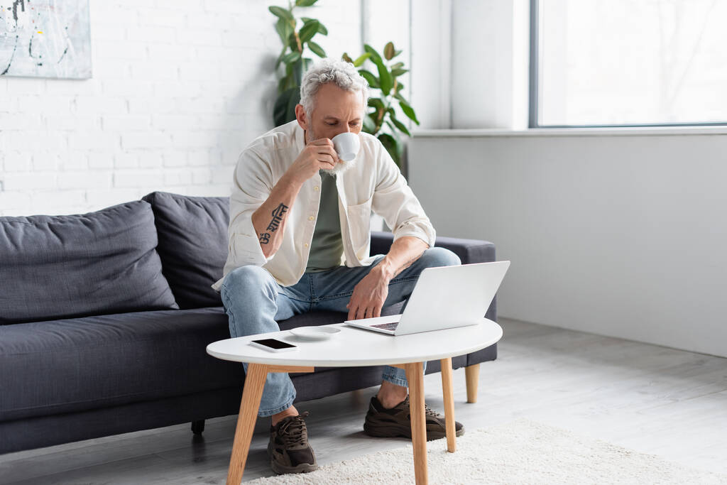 bearded man drinking coffee and using laptop near smartphone with blank screen on coffee table - Photo, Image