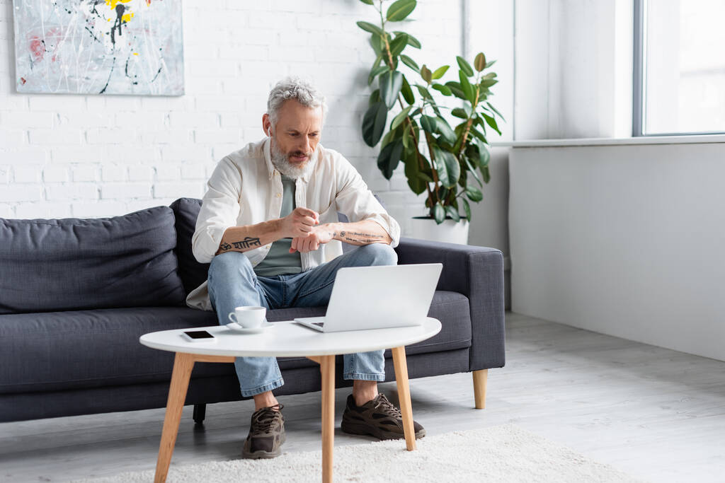 szakállas férfi mutató laptop, miközben ül a kanapén közel csésze kávé és okostelefon üres képernyő dohányzóasztalon - Fotó, kép