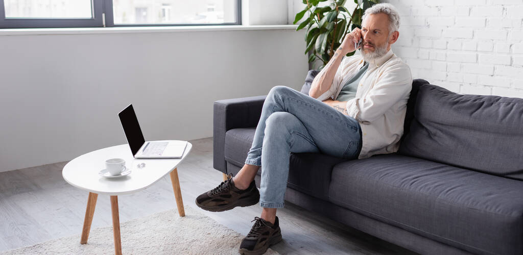hombre barbudo hablando en el teléfono inteligente mientras está sentado en el sofá cerca de la computadora portátil con pantalla en blanco, pancarta - Foto, imagen