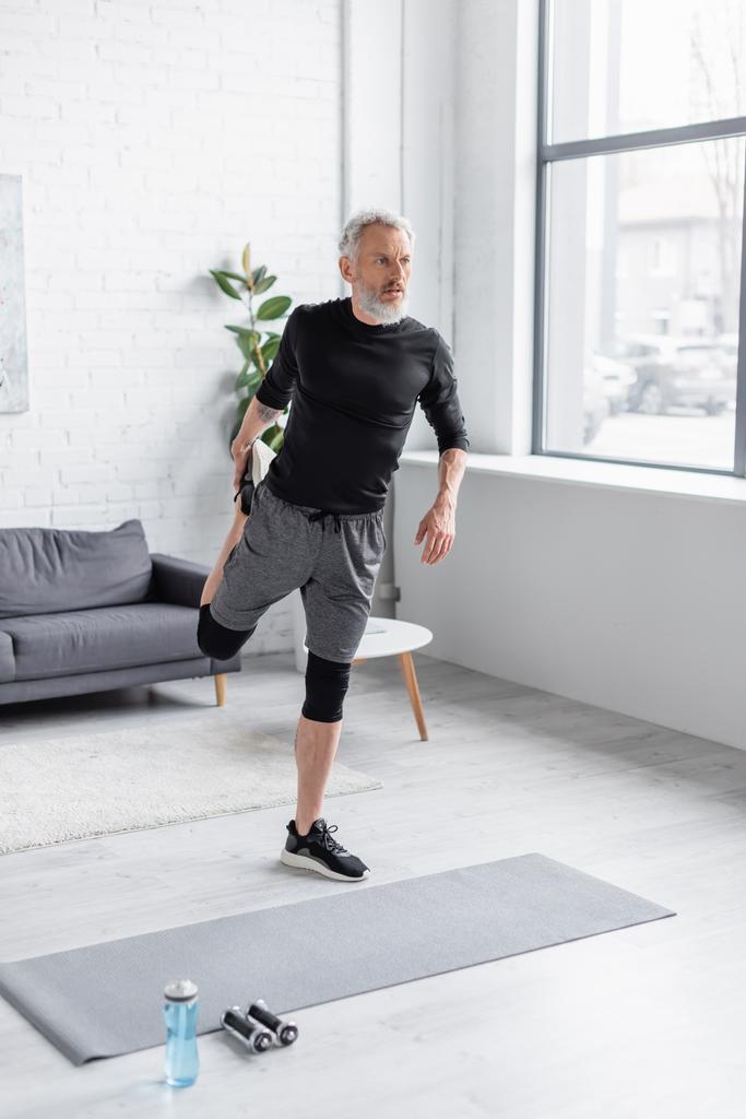 Vollbärtiger und tätowierter Mann in Sportbekleidung beim Training auf Fitnessmatte in der Nähe von Hanteln im Wohnzimmer  - Foto, Bild