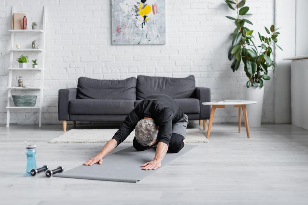 homem com cabelos grisalhos esticando no tapete de fitness perto de halteres na sala de estar, banner - Foto, Imagem