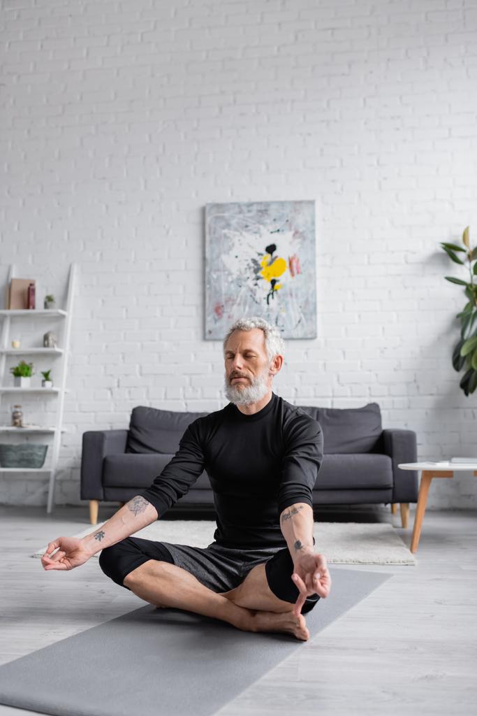 bärtiger Mann mit geschlossenen Augen meditiert auf Yogamatte neben Hanteln  - Foto, Bild